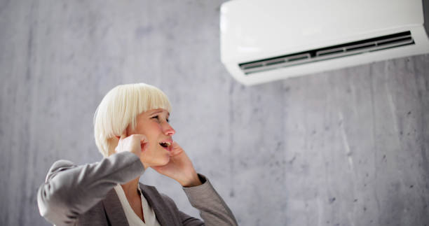 A woman observes an air conditioner, which is producing a loud noise, as she considers its impact on her environment.