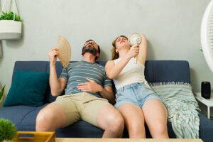 A man and woman sit on a couch, looking uncomfortable as a fan blows air, with the AC not cooling the room.

