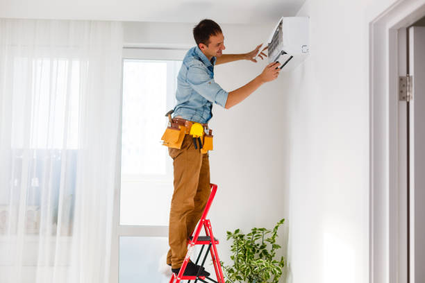 A man using a ladder to fix an AC unit, demonstrating maintenance work on a warm day.