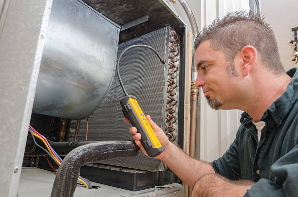 A man wearing a blue coat is engaged in air conditioner repair, specifically checking for refrigerant leaks.