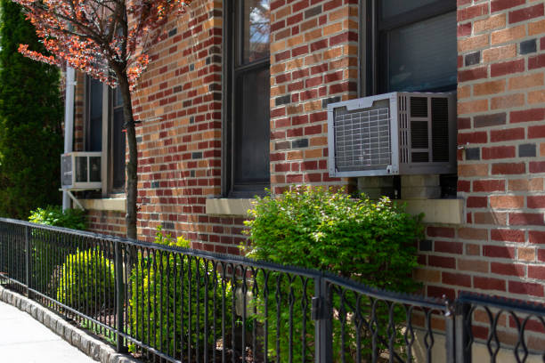 Outdoor window air conditioning units on an old New York City brick apartment building with green plants along a sidewalk