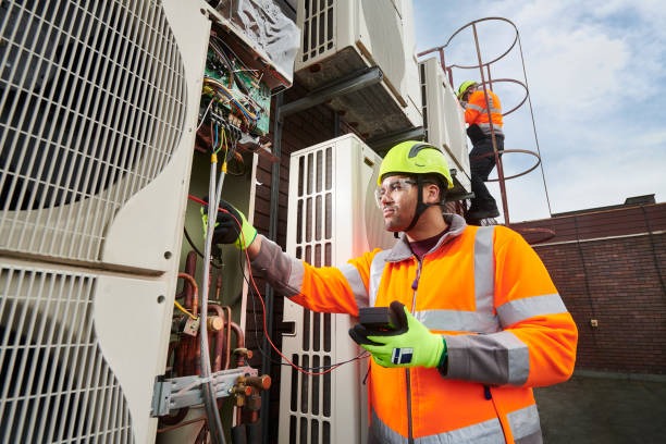 A man in an orange safety vest and hard hat repairs an air conditioner unit on a rooftop.

