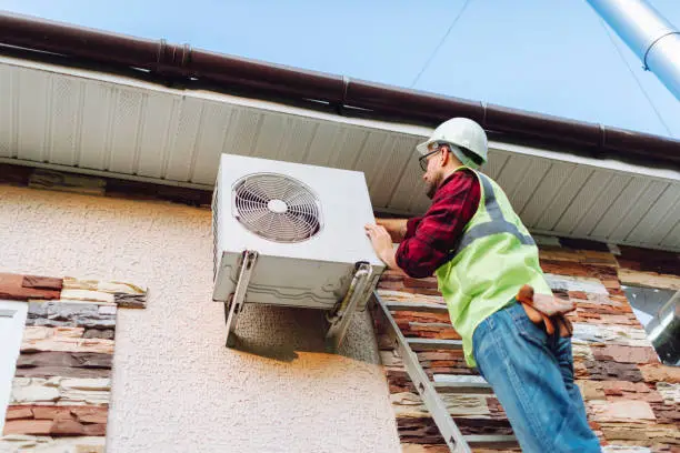 A man in a hard hat and safety vest is replacing an air conditioner on a building rooftop.